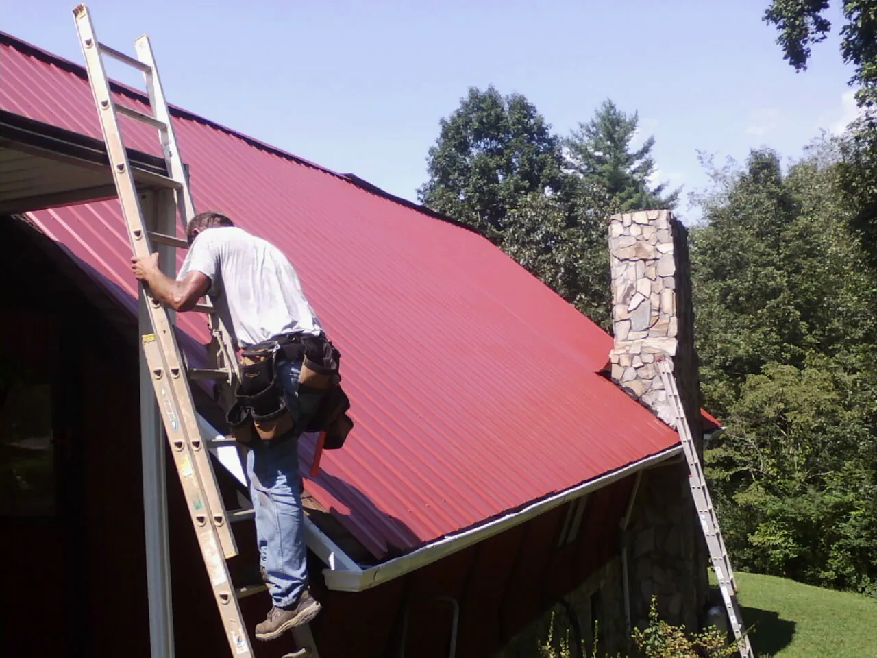 Roofer on a ladder beside a red metal roof and stone chimney — Soffit & Fascia Repair by Roofing Brothers in Zelienople